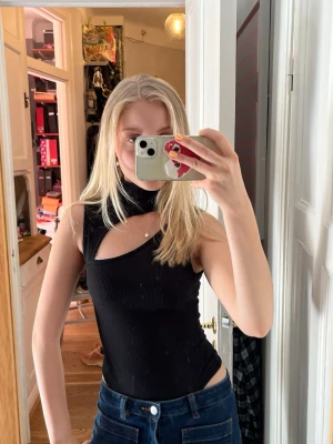 Svart asymmetrisk ribbad body  - Säljer en svart ribbad body med asymmetrisk design och hög hals. Toppen är ärmlös och har en cool cut-out detalj fram och bak