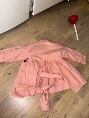 Rosa trenchcoat med knytskärp - Snygg rosa trenchcoat med dubbelknäppning och stora bruna knappar. Jackan har ett klassiskt knytskärp i midjan och vida ärmar med veckdetaljer. Tillverkad i ett lätt bomullsliknande material och har en avslappnad, något utsvängd passform.