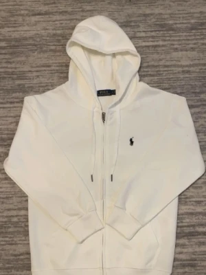 Vit Polo Ralph Lauren hoodie - Säljer en stilren vit hoodie från Polo Ralph Lauren med dragkedja och klassisk huva. Tröjan har det ikoniska mörkblå broderade logotypen på bröstet och justerbara snören i huvan. Perfekt för en clean och avslappnad look.
