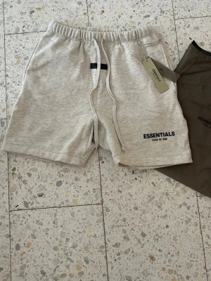 Grå shorts från Essentials Fear of God - Snygga ljusgrå shorts från Essentials Fear of God med broderad logga på benet och svart patch på framsidan. Shortsen har snörning i midjan och är tillverkade i mjuk bomull, perfekta för chill dagar eller träning.