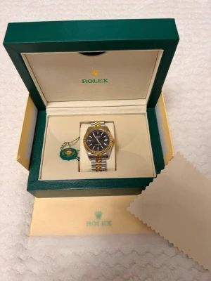  guld och silver klocka - Säljer en klassisk med svart urtavla, guldfärgad räfflad bezel och tvåtonad länk i guld och silver. Klockan kommer i originalbox med certifikat, extra länkar och verktyg. Perfekt accessoar för dig som gillar exklusiv stil.