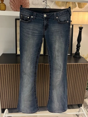 Reserverad  - Den här varan är reserverad! Säljer ett par mörkblå bootcut jeans från True Religion med klassiska kontrastsömmar och broderade bakfickor. Jeansen har låg midja, fem fickor och silverfärgade knappar. Perfekta för en avslappnad men snygg stil. Använda cirka 3 gånger. Relativt stretchiga. Midja: 41 cm. Lårvidd: 24 cm. Innerbenslängd: 80 cm. Fotvidd: 27 cm. 