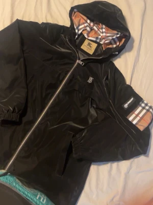 Svart Burberry vindjacka med rutigt foder - Snygg svart vindjacka från Burberry med klassiskt rutigt foder i huvan och på ärmen. Jackan har dragkedja framtill, elastiska muddar och fickor. Burberry-logga på bröstet och ärmen ger en exklusiv känsla. Perfekt för dig som vill ha en stilren och trendig look.