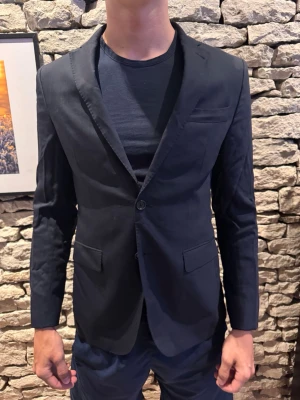 Svart klassisk kavaj med två knappar - Stilren svart kavaj med smal passform och två knappar framtill. Kavajen har två fickor med lock och en bröstficka. Perfekt till både t-shirt och skjorta för en snygg look. Materialet ser ut att vara en blandning av bomull och polyester.