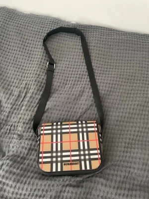 Burberry axelväska med rutigt mönster - Snygg axelväska från Burberry med klassiskt beige, svart, vitt och rött rutmönster på framsidan och svart baksida. Justerbar svart axelrem och stilren fyrkantig form. Perfekt för dig som vill ha en ikonisk och trendig accessoar. Har aldrig använt den.