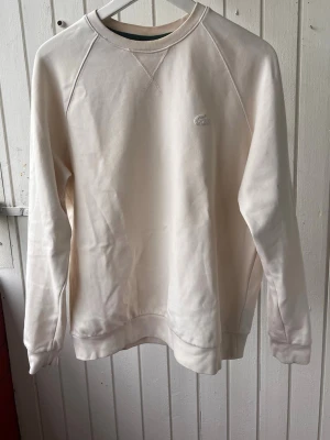 Vit sweatshirt från Lacoste - Klassisk vit sweatshirt från Lacoste med rund halsringning och diskret broderad logotyp på bröstet. Tröjan har långa ärmar, ribbade muddar och är tillverkad i mjukt bomullsmaterial. Perfekt för en clean och stilren look. Inga fläckot osv