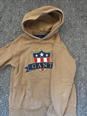 Beige hoodie från GANT med tryck - Snygg beige hoodie från GANT med stort tryck på bröstet i form av ett sköldmärke med stjärnor och ränder i blått, vitt och rött. Hoodien har huva och en klassisk känguruficka framtill. Tillverkad i mjukt bomullsmaterial för skön känsla.