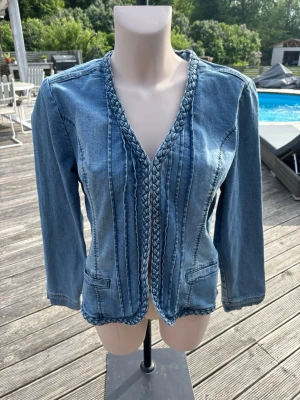 Ljusblå jeansjacka med flätade detaljer - Snygg ljusblå jeansjacka med unika flätade detaljer längs krage och framkant. Jackan har lång ärm, två dekorativa fickor framtill och en figurnära passform. Perfekt för dig som vill ha en lite annorlunda jeansjacka med coola detaljer.
