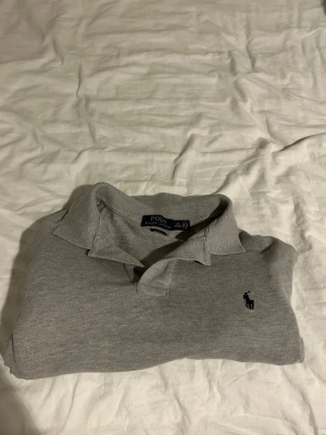 Grå långärmad piké från Polo Ralph Lauren - Klassisk grå långärmad pikétröja från Polo Ralph Lauren med mörkblå broderad logga på bröstet. Skjortan har krage och knappar vid halsen. Perfekt för en clean och stilren look.                                  . Jag köpte tröjan av mammas vän för 500kr förra året misstag för den var för liten för mig knappt använd men en liten fettfläck vilket försvinner om man använder medel. Som sagt jag har inte använt tröjan därför har jag inte tvättat den något.