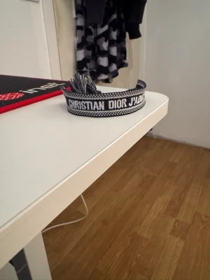Christian Dior J'Adior armband - Snyggt vävt armband från Christian Dior med texten 'J'Adior' i vitt på svart bakgrund. Armbandet har justerbar knytning med fransar i ändarna och en cool, modern look. Perfekt accessoar för att lyfta din outfit med en exklusiv touch.