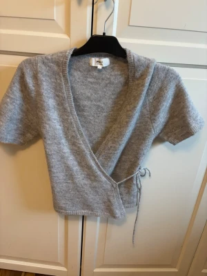 Grå omlottopp i ullmix Gina Tricot - Kortärmad stickad omlottopp från Gina Tricot i mjuk grå ullmix. Tröjan har knytband i sidan och djup v-ringning. Perfekt för lager på lager och en trendig look.
