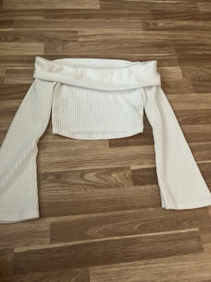Vit offshoulder ribbad topp - Trendig vit offshoulder topp med ribbad struktur och långa ärmar. Toppen har en croppad passform och är perfekt för dig som gillar en stilren och modern look. Materialet känns mjukt och stretchigt, vilket gör den bekväm att bära.