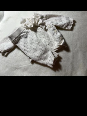 Vitt stickat babyset med volang - Sött tvådelat babyset i vitt med små prickar, volangkrage och knappar på tröjan. Setet består av långärmad tröja och byxor med ribbade muddar och dekorativ kant. Mjukt stickat material, perfekt för mysiga dagar.