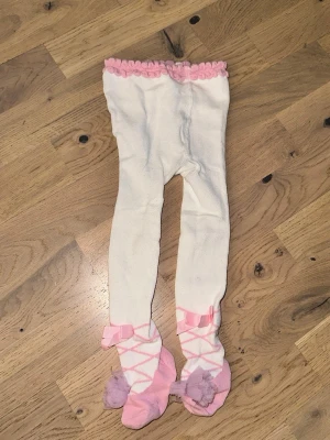 Vita tights med rosa detaljer - Söta vita tights med rosa volangkant upptill och ballerina-inspirerade mönster på fötterna. Tightsen har rosa rosetter och lila pompoms på fötterna, vilket ger en lekfull look. Materialet är mjukt och stretchigt, perfekt för att sitta bekvämt hela dagen.