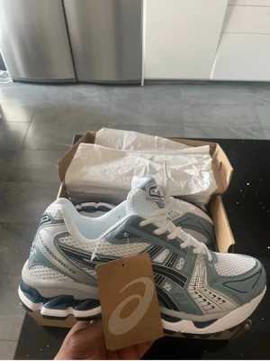 Asics Gel-Kayano 14 vita och silver - Säljer ett par Asics Gel-Kayano 14 sneakers i vitt och silver med svarta detaljer. Skorna har en sportig design med mesh- och syntetmaterial för bra andningsförmåga och komfort. Perfekta för dig som gillar retrostil och streetwear.
