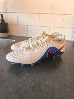 Nike vita och blå fotbollsskor - Säljer ett par stilrena fotbollsskor från Nike i vitt och blått med rosa detaljer. Skorna har en låg profil, snörning och en yttersula med fasta dobbar för bra grepp på planen. Materialet är syntetiskt och designen är modern med en stor Nike-logga på sidan. 