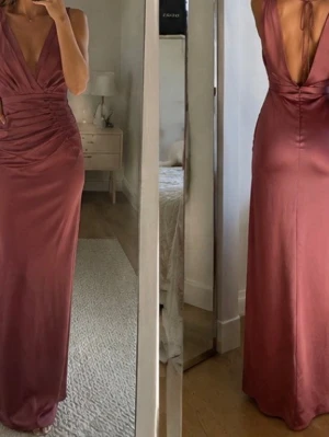 Rosa långklänning med drapering - Säljer en elegant rosa långklänning med djup v-ringning och snygga draperade detaljer framtill. Klänningen har knyt i nacken och är ärmlös, vilket ger en stilren och modern look. Perfekt för dig som vill sticka ut med en unik och feminin stil.