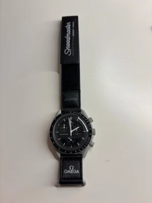 Omega x Swatch Speedmaster klocka - Snygg svart Omega x Swatch Speedmaster klocka med svart textilarmband och tydliga loggor på bandet. Klockan har en klassisk rund urtavla med kronograf och vita detaljer. Baksidan har en måndekor och text. Perfekt för dig som gillar ikoniska samarbeten.