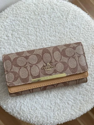 Coach brun monogram plånbok - Snygg plånbok från Coach i brunt monogramtyg med klassiskt C-mönster. Plånboken har guldfärgade detaljer och en beige kant, samt Coach-logga i metall framtill. Perfekt för dig som gillar stilrena accessoarer med lyxig känsla.