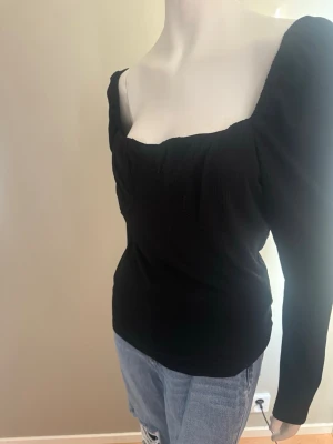 Svart långärmad topp med fyrkantig urringning strl XL - Trendig svart topp med lång ärm och snygg fyrkantig urringning. Toppen har figurnära passform och lätt puffade axlar som ger en modern siluett. Tillverkad i ett ribbat material som känns mjukt mot huden. Perfekt att styla med jeans eller kjol.