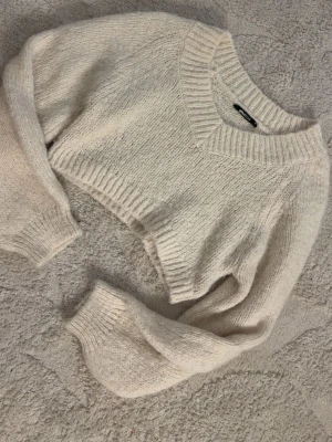 Vit cropped stickad tröja Gina Tricot - Säljer en vit cropped stickad tröja från Gina Tricot. Tröjan har v-ringning, bred ribbad kant vid halsen och långa, lite pösiga ärmar med ribbade muddar. Perfekt för lager på lager och en chill look.
