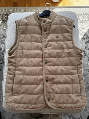 Beige manchester-dunväst från Morris - Stilren beige dunväst från Morris i mjuk manchester. Västen har tryckknappar framtill och diskret logga på bröstet.