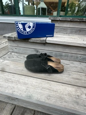 Svarta Birkenstock tofflor mocka - Säljer ett par svarta Birkenstock tofflor i mocka med klassisk design och justerbart spänne. De har en bekväm fotbädd i kork och öppen häl, perfekta för chill dagar hemma eller ute. Stilen är tidlös och passar till det mesta.