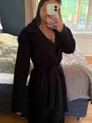 Svart lång kappa med knytskärp - Stilren svart kappa i lång modell med breda slag och knytskärp i midjan. Klassisk design utan synliga knappar framtill och raka ärmar. Perfekt för dig som gillar en enkel men trendig look. Materialet ser ut att vara mjukt och något tjockare, passar bra till höst och vinter.