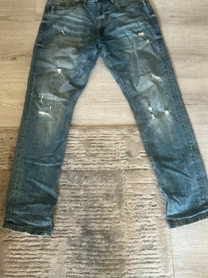 Blå slitna jeans  - Säljer ett par blå jeans med slitningar och distressed detaljer på benen. Klassisk femficksmodell med ljus tvätt och synliga sömmar. Perfekta för en avslappnad och trendig look.