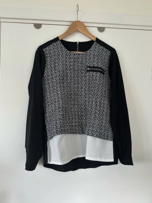 Svart och vit blus med kontrasttyg, storlek M - Fin svart och vit blus med långa ärmar i storlek M. Blusen har en mönstrad framsida i svart och vitt i stickat kontrasttyg, med en liten dragkedjeförsedd ficka på bröstet. Baksidan och ärmarna är helsvarta. Nedtill har blusen ett vitt lager som sticker ut under det mönstrade partiet. Dragkedja baktill i nacken. Mycket fint skick, som ny! 