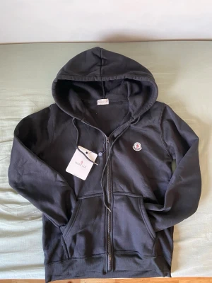 Svart hoodie med dragkedja från Moncler - Svart hoodie från Moncler med dragkedja framtill, justerbar huva med snören och två fickor. Klassisk Moncler-logga på bröstet. Tillverkad i mjukt bomullsmaterial som känns skönt mot huden. Perfekt för en avslappnad och stilren look.