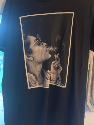 Svart t-shirt med tryck från L'Arte - Cool svart t-shirt från L'Arte med ett stort svartvitt tryck på framsidan. T-shirten har klassisk passform och rund halsringning. Perfekt för dig som gillar streetstyle och vill sticka ut med en unik printad design.