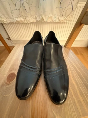 Svarta lackloafers från MoodA - Snygga svarta loafers i lack från MoodA med klassisk design och diskreta sömdetaljer framtill. Skorna har en stilren form och blanka ytor som ger en elegant look. Perfekta för dig som vill ha en dressad stil med lite edge.