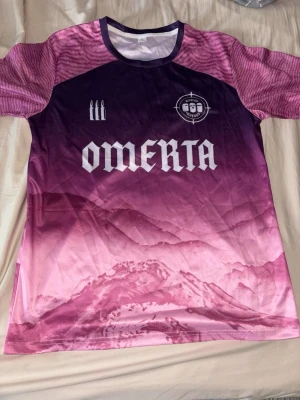 Lila och rosa Omerta t-shirt - Säljer en unik t-shirt från Omerta med snyggt bergsmotiv i lila och rosa toner. T-shirten har korta ärmar, rund halsringning och tryckt logga samt text framtill. Perfekt för dig som vill sticka ut med cool design.