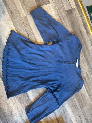 Blå ribbad topp från 157 - Säljer en blå ribbad topp från 157 i storlek XS. Toppen har knappar framtill och dekorativa volangdetaljer vid ärmslut och nedtill. I strl XS men passar även S. Den är nästan aldrig använd och i väldigt bra skick,kontakta mig vid frågor.