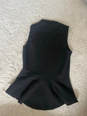 Svart ärmlös peplumtop från H&M - Snygg svart ärmlös peplumtop från H&M med hög hals. Toppen har en figurnära passform upptill och en utställd volang nedtill.