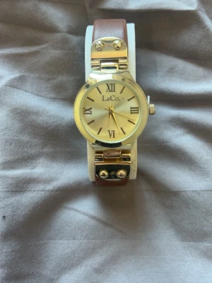 Guldig klocka från La&Co med läderband - Stilren klocka från La&Co med rund guldfärgad boett och romerska siffror på urtavlan. Klockan har ett brunt läderband och detaljer i guld, inklusive dekorativa skruvar på bandfästet. Perfekt accessoar för en snygg look.