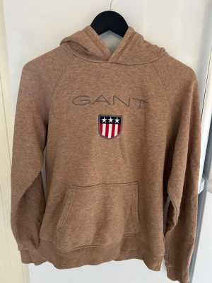 Beun hoodie från GANT med logga - Snygg brun hoodie från GANT med broderad logga och sköld på bröstet. Klassisk känguruficka framtill och huva. Perfekt för en avslappnad och stilren look. Mjuk bomullsblandning som känns skön mot huden.