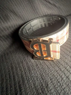 Beige och rutig Burberry-inspirerat bälte - Snyggt bälte med klassiskt beige och rött rutmönster samt en stor guldfärgad B-spänne framtill. Bältet har svarta detaljer på insidan och passar perfekt för att lyfta din outfit.