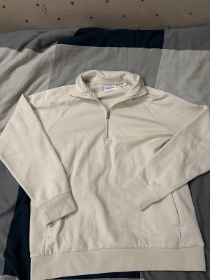 Beige half zip hoodie från Lindbergh - Snygg beige hoodie från Lindbergh med half zip-dragkedja och broderad logga på bröstet. Tillverkad i mjuk bomull och har en hög krage för extra stilpoäng. Perfekt för dig som gillar en clean och enkel look.