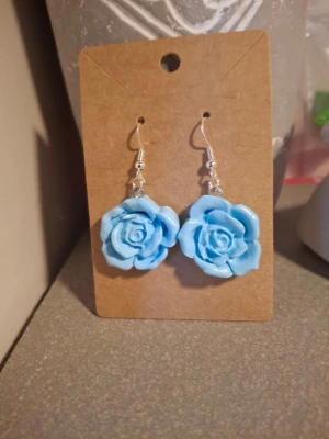 Blå rosformade örhängen - Säljer ett par unika örhängen formade som blå rosor. De har krokar i silver och själva rosorna är gjorda handgjorda i lera.. Perfekta för dig som vill sticka ut med en färgglad accessoar.