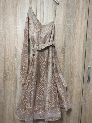 Champagnefärgad glittrig klänning - Snygg champagnefärgad klänning med glittriga detaljer och bälte i midjan. Klänningen har ett transparent lager med broderat mönster, en lång ärm och en axelbandslös sida för en unik oneshoulder-look. Perfekt för dig som vill sticka ut. Har en extra arm ifall man vill, köpte den utomlands och är använt 1 gång och fortfarande nu. Perfekt för fester, bröllop och allt möjligt.