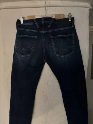 Mörkblå Replay jeans med smal passform - Snygga mörkblå jeans från Replay med smal passform och klassiska fem fickor. Jeansen har slitningar och ljusare partier framtill samt dekorativa sömmar på bakfickorna. Tillverkade i denim med stretch för extra komfort.
