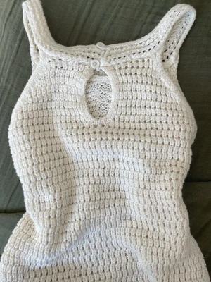 Vit virkad sommartopp - Supersöt virkad topp i vitt med breda axelband och en liten knappdetalj i nacken. Toppen har ett hålformat mönster framtill och är figurnära med stretchig passform. Perfekt för varma dagar och enkel att styla till jeans eller kjol.