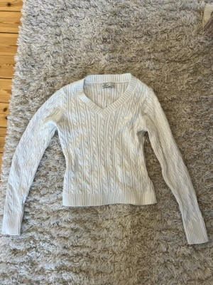 Vit kabelstickad tröja Polo Ralph Lauren - Kabelstickad vit tröja från Polo Ralph Lauren med V-ringning och långa ärmar. Tröjan har ribbade muddar vid ärmslut och nederkant samt den klassiska broderade loggan på bröstet. Perfekt för dig som gillar en clean och tidlös stil.