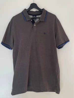 Champion piké – grå – L - Grå pikétröja från Champion med klassisk passform och diskret logga på bröstet. Stilren modell med blå detaljer vid krage och ärmslut.  ✔ Storlek: L ✔ Färg: Grå ✔ Modell: Piké ✔ Skick: Bra skick