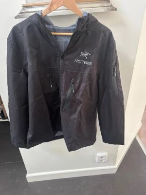 Svart vindjacka från Arc'teryx - Svart vindjacka från Arc'teryx med huva och flera dragkedjeförsedda fickor. Jackan har logga på bröstet och en sportig look. Perfekt för blåsiga dagar och outdoor-aktiviteter.