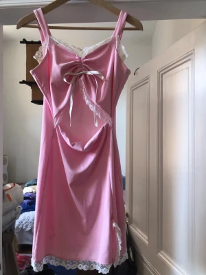 Rosa ribbad kortklänning med spets - Supersöt rosa kortklänning med ribbad struktur, spetskant vid urringning och nederkant samt en dekorativ rosett framtill. Klänningen är ärmlös med breda axelband och har omlottdetalj vid bysten. Ärvd klänning så är osäker på märket men skulle gissa på Shein…Perfekt för sommarens alla tillfällen. Passar både SX/S/M!Finns små missfärgningar som jag kan skicka om så önskas💐🌷🌸