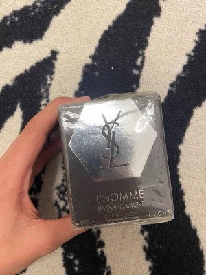 Yves Saint Laurent L'Homme EdT 60ml - Yves Saint Laurent L'Homme Eau de Toilette 60ml i elegant grå och silverfärgad förpackning. Parfymen har en modern, maskulin design med YSL-logga på framsidan. Flaskan är tillverkad av glas och förpackningen är plastad för extra skydd.