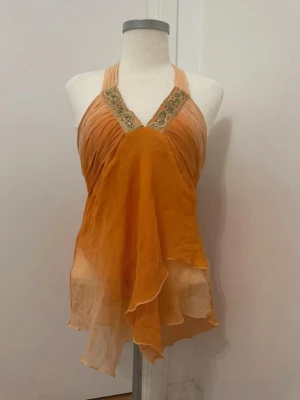 Orange asymmetrisk halterneck-topp i 100% silke - Säljer en unik halterneck-topp i orange/korall med knyt i nacken och spetsdetaljer och paljetter vid v-ringningen. Toppen har ett asymmetriskt, flödande tyg i silke som ger en bohemisk vibe. Perfekt för dig som vill sticka ut med en cool och luftig sommartopp.